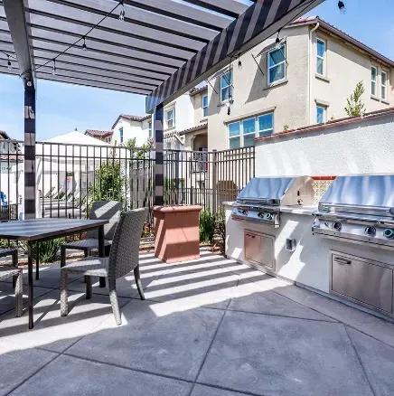 Patio with outdoor dining table, lounge chair, and stainless steel grill under a pergola in a sunny courtyard