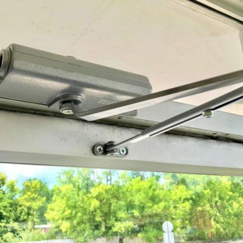Door Closers Installation and Repair - A close up of a door closer on a window with trees in the background Door Closers Installation and Repair - A close up of a door closer on a window with trees in the background