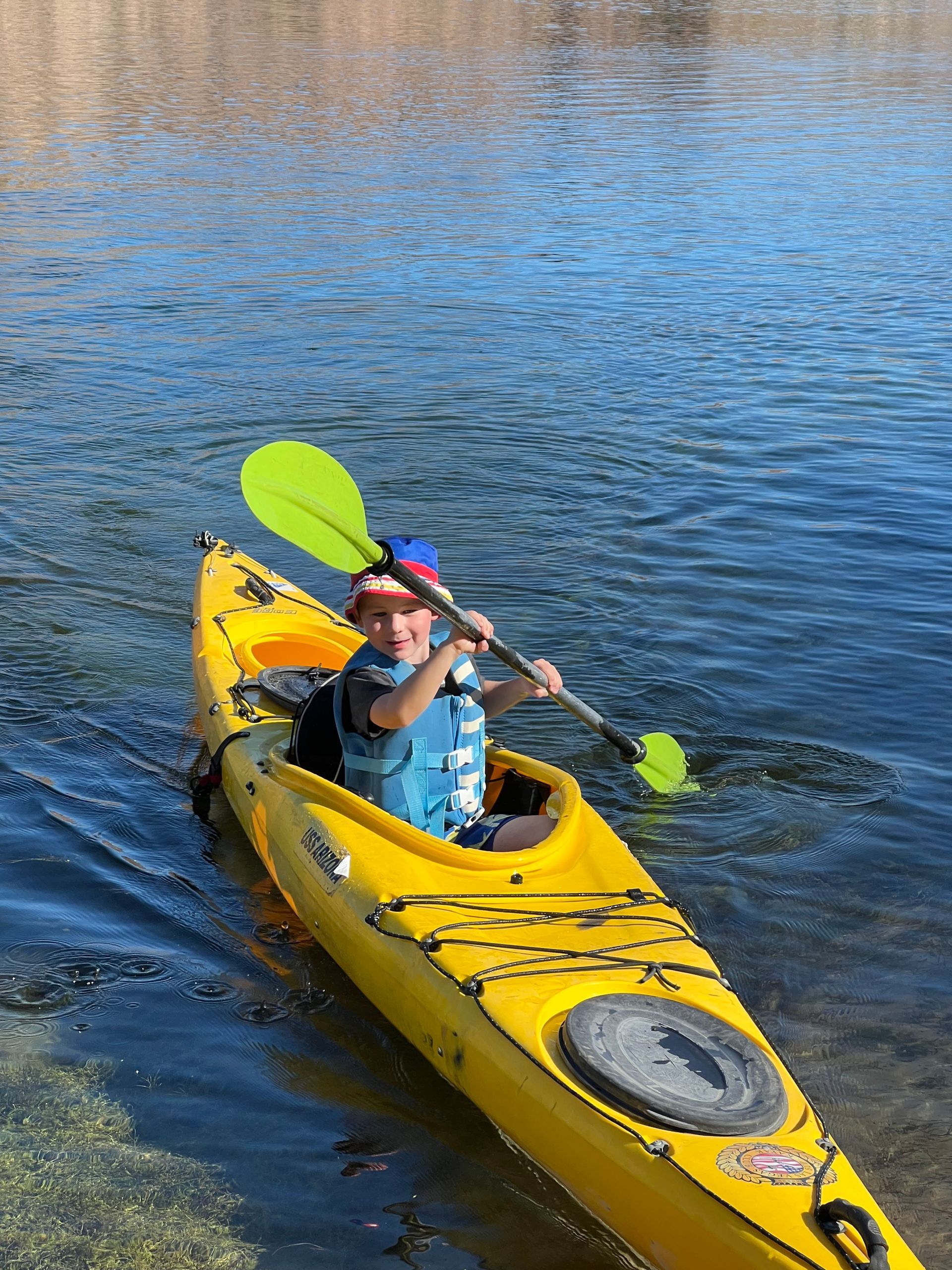 Person in yellow kayak paddles on a blue lake, wearing a life vest and a hat.