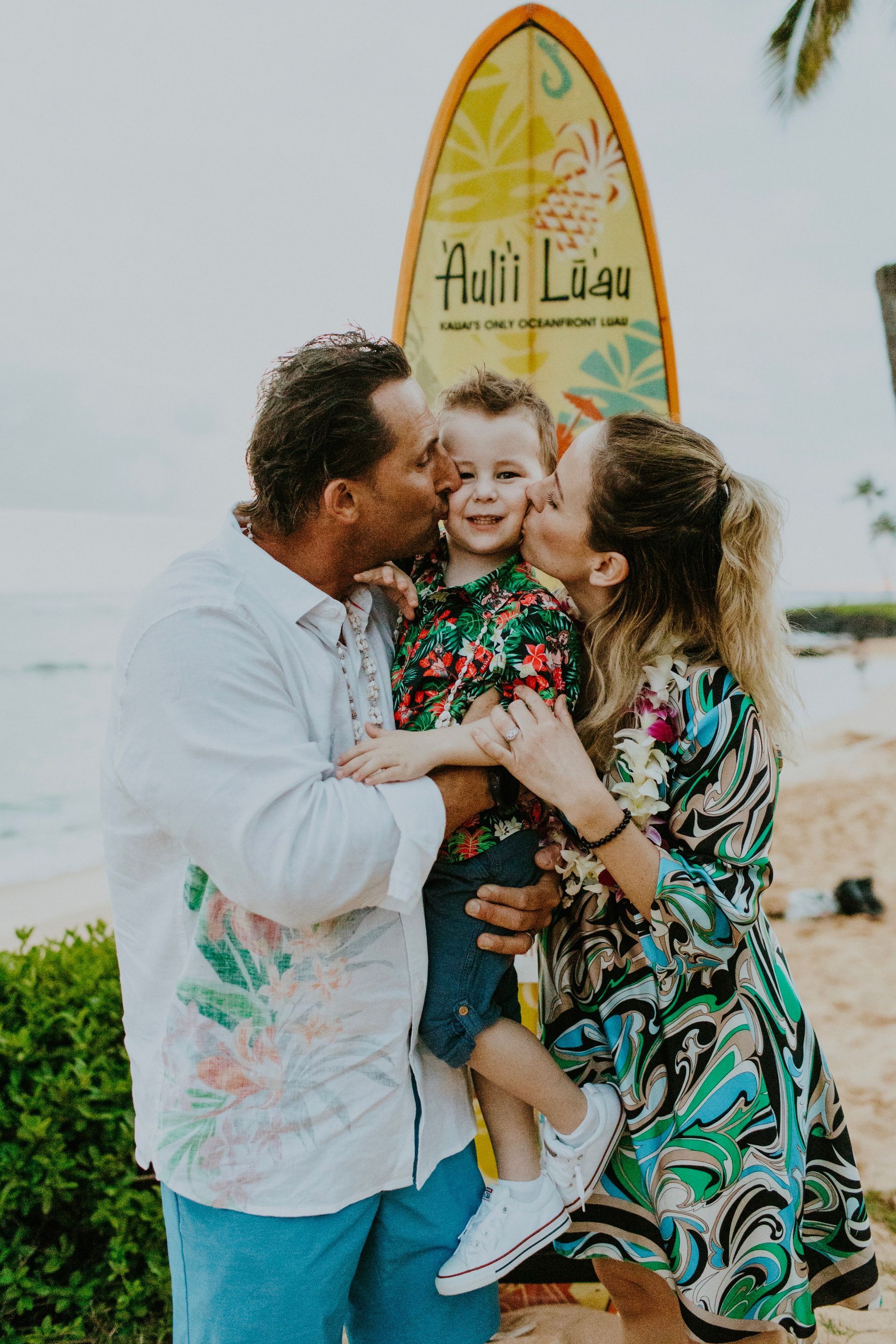 Family kissing a child on cheeks in front of a surfboard on a beach.