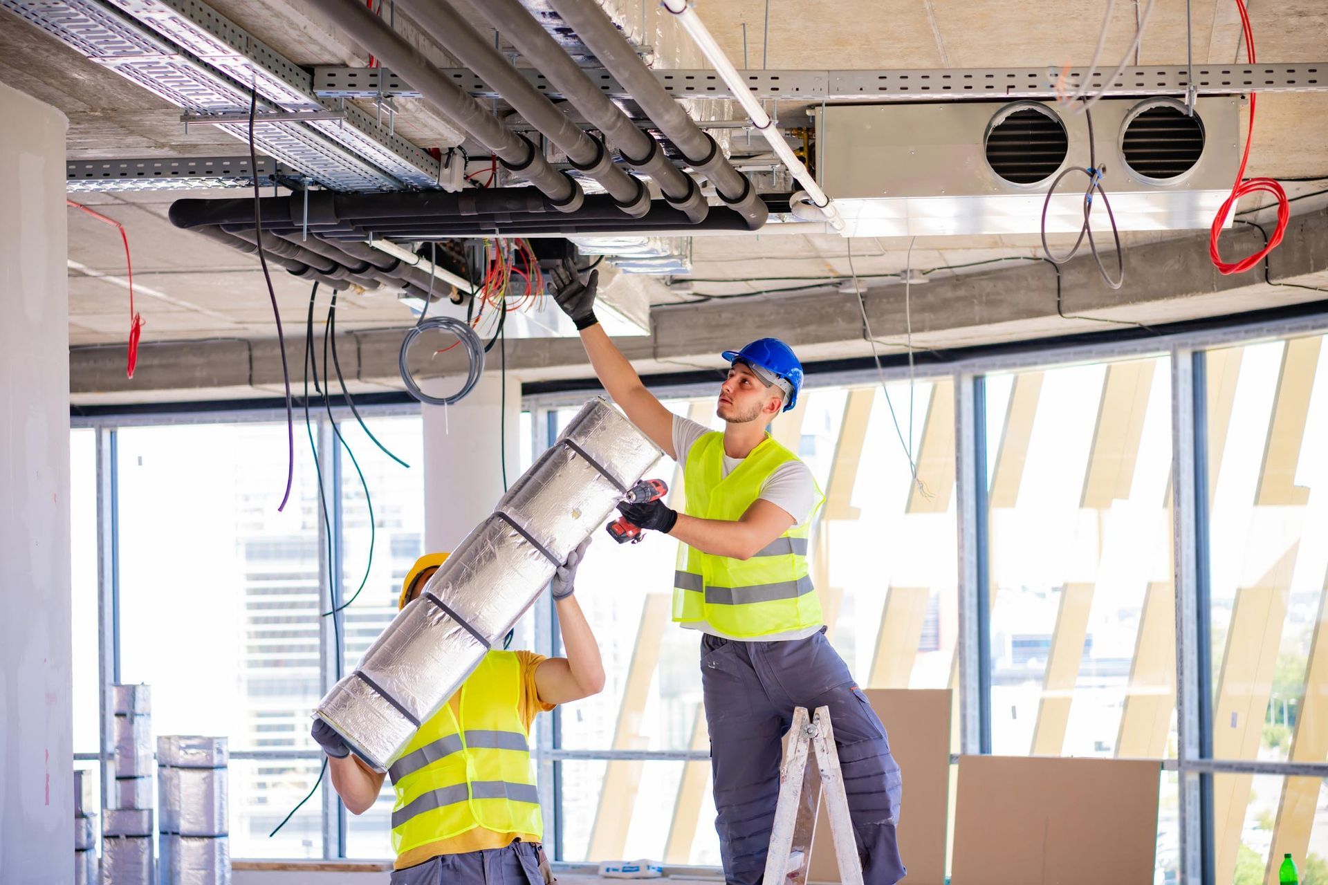 Two construction workers installing ventilation in a building under construction, wearing safety vests and helmets.
