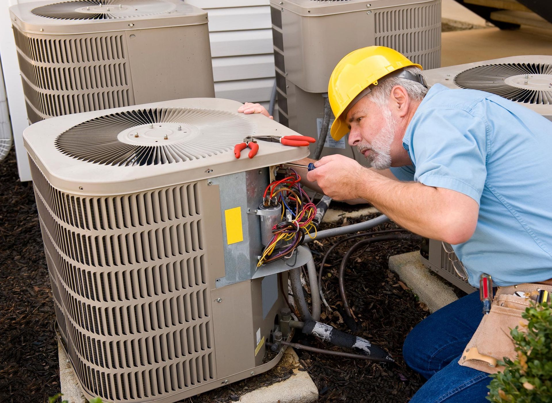HVAC technician in yellow hard hat repairing an air conditioner unit outdoors.