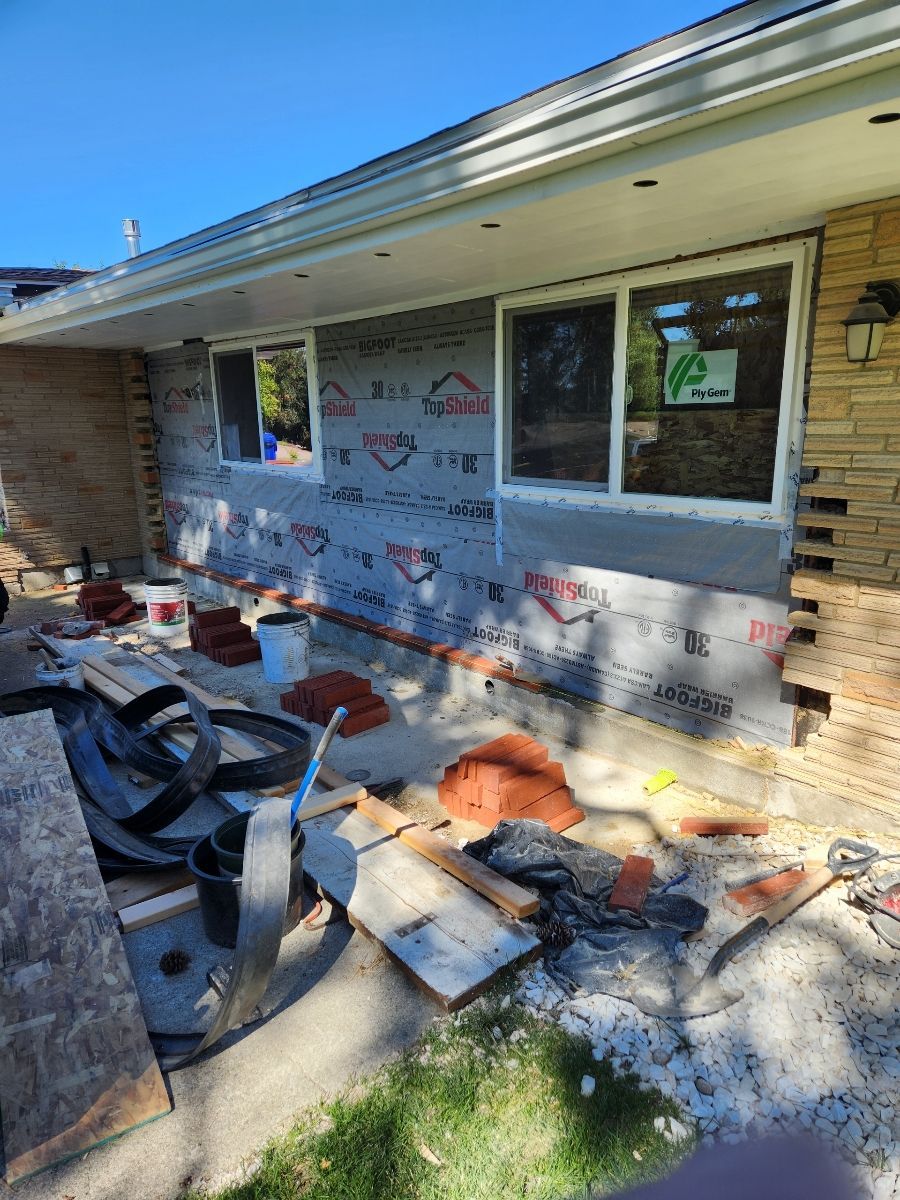 Construction site: brick home exterior, new windows, covered with protective material, tools and debris on ground.