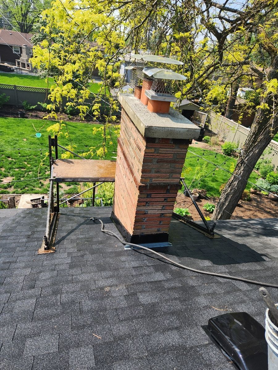 Brick chimney on a shingled roof, with scaffolding next to it; green lawn and trees in background.