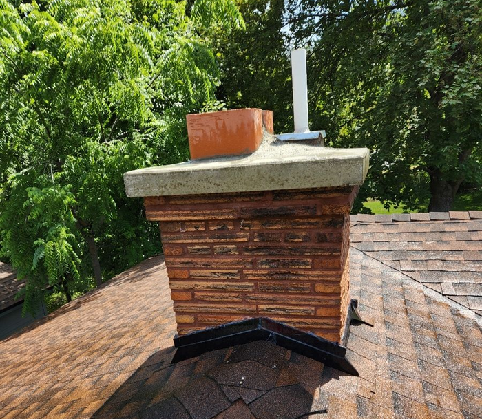 Brick chimney on a brown shingled roof, topped with a concrete cap and a flue. Green trees in background.