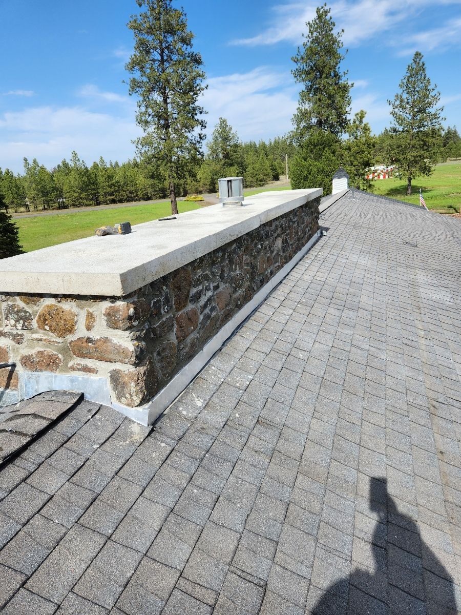 Stone chimney with concrete cap on a shingle roof against a green field and blue sky.