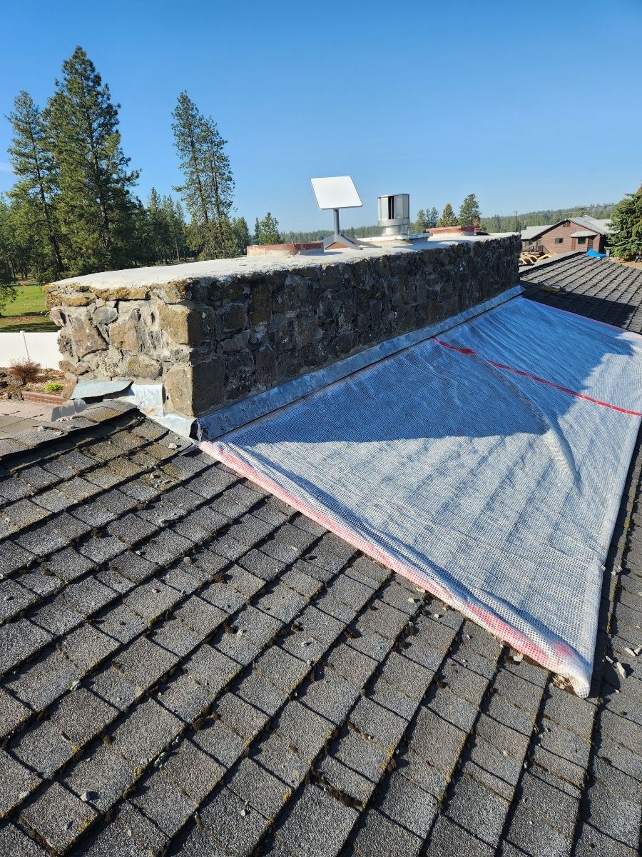 Roof with chimney, partially covered with white material. Sky, trees in background.