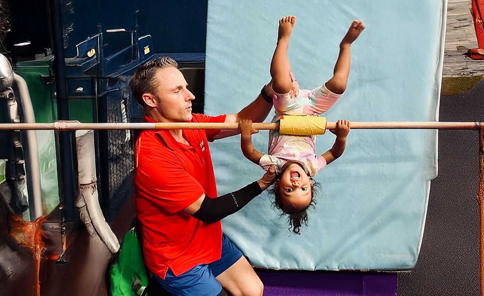 Man helps a smiling child hanging upside down on a bar. Red shirt, blue shorts. Gym setting.