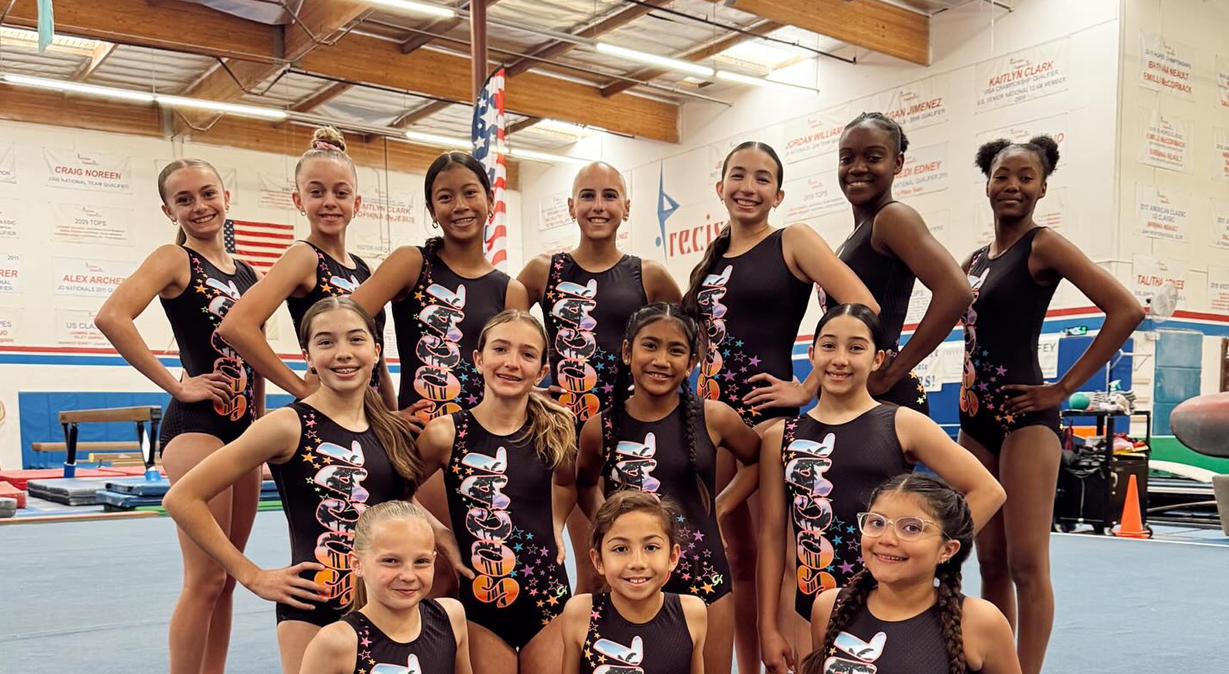 Gymnastics team poses in matching black leotards, hands on hips, in a gym.