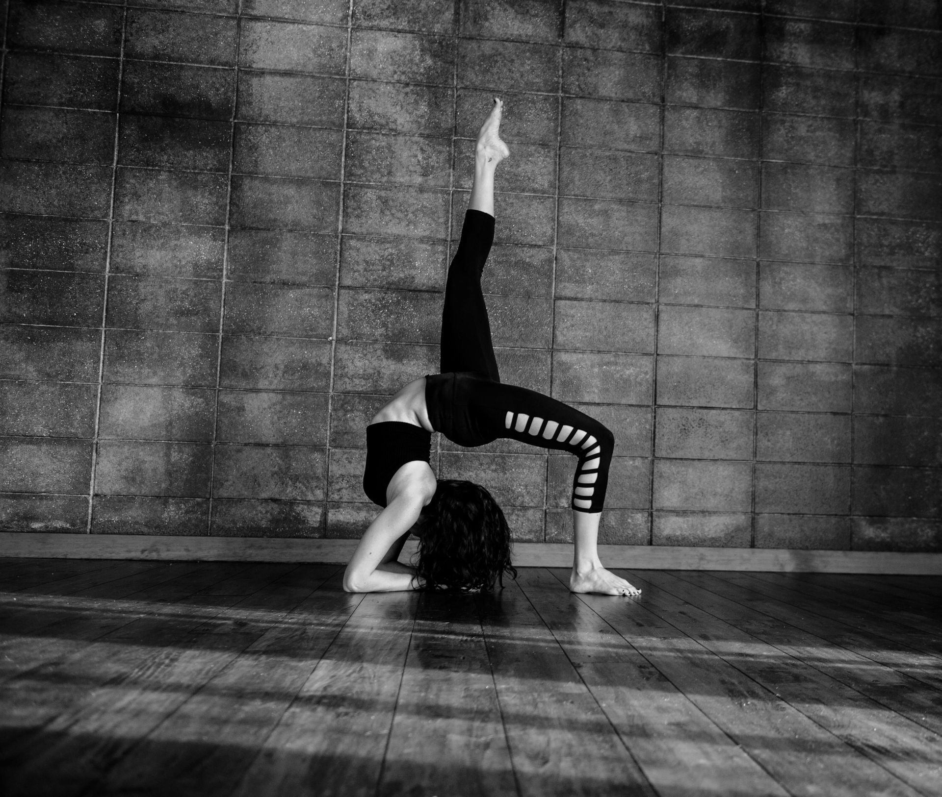 Woman in a bridge pose, one leg extended, against a textured wall and wooden floor. Black and white.