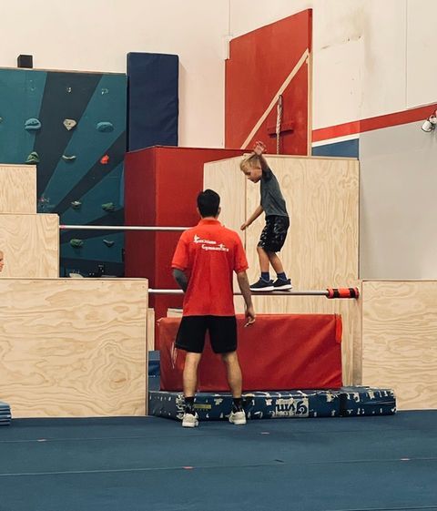 A child balances on a bar with an instructor nearby. They're in a gym with obstacles.