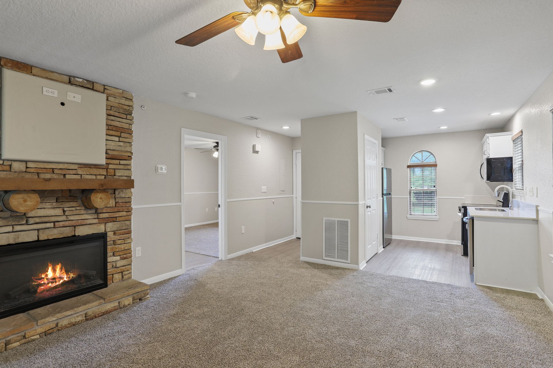 Photo of a living room with a fireplace and ceiling fan, and the hallway, a bedroom, and the kitchen in the background