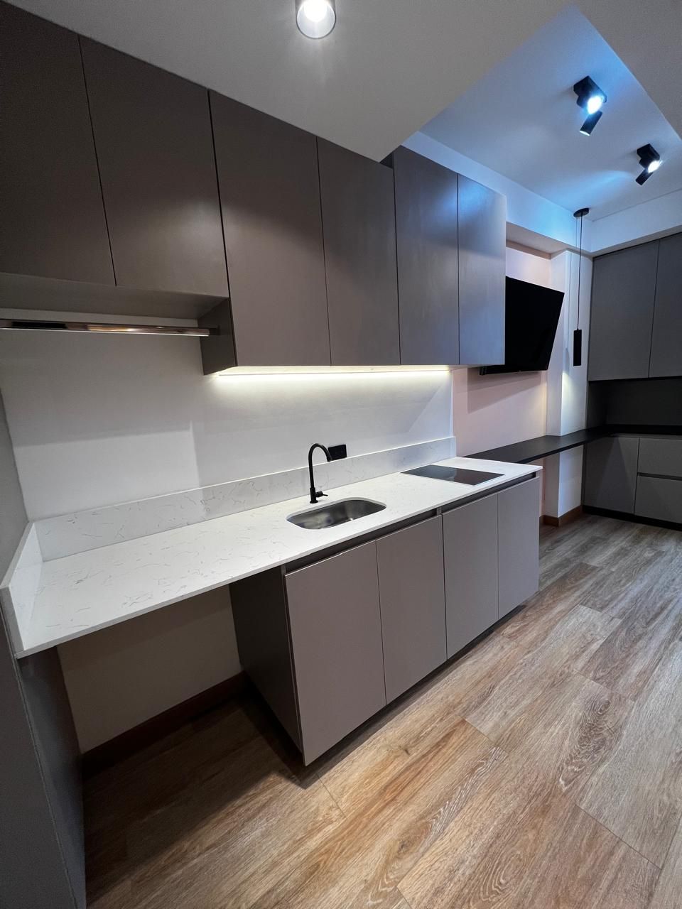 Cocina moderna con gabinetes grises, encimeras blancas, fregadero, estufa y piso de madera bajo luces empotradas.