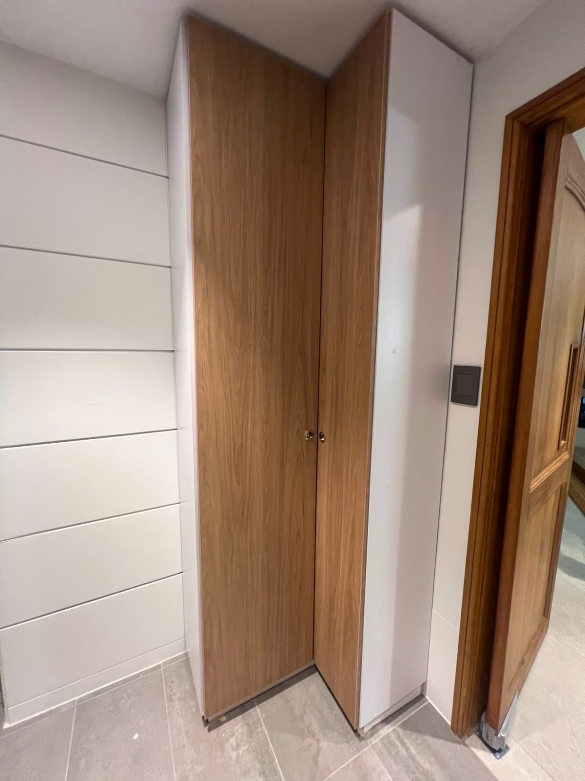 Puerta de un cubículo de baño con paneles de madera junto a paredes de azulejos blancos y un marco de puerta de madera.