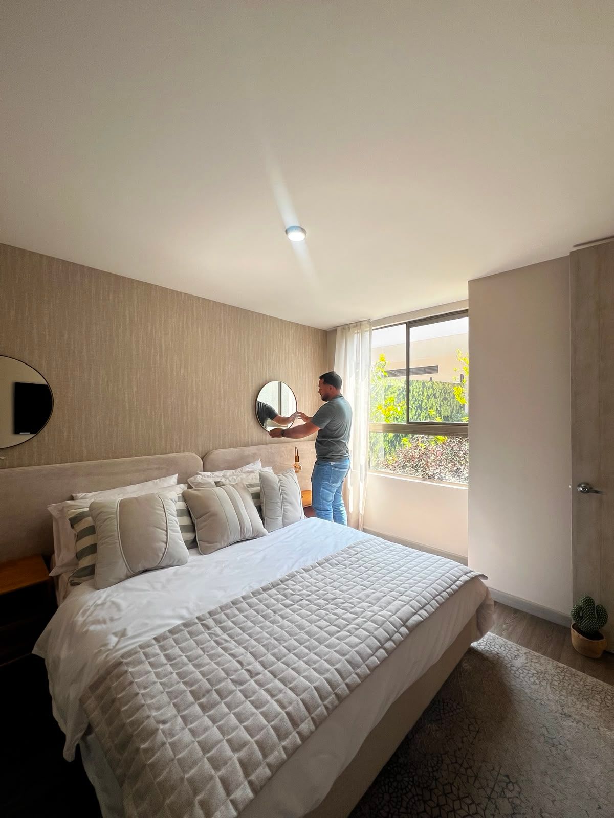 Habitación luminosa con la cama hecha; una persona está de pie junto a la ventana mirando hacia afuera.