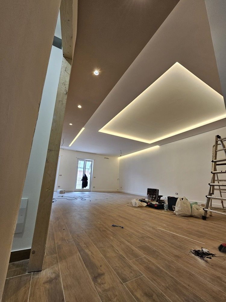Habitación moderna vacía en proceso de renovación con suelo de madera, luces de techo inclinadas y una escalera a la derecha.