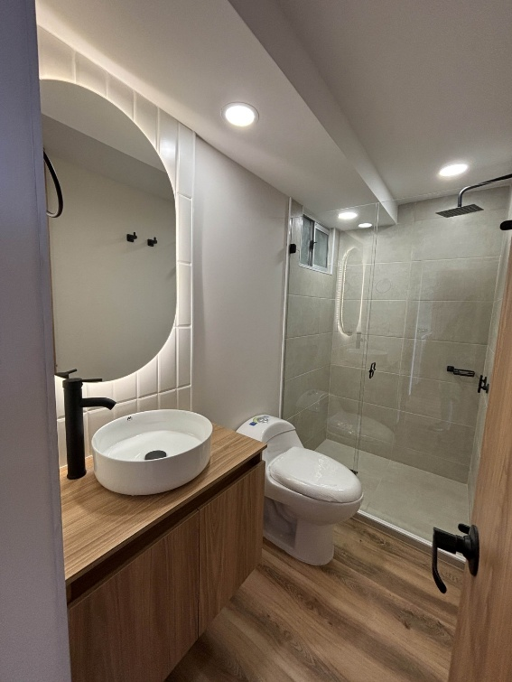 Baño moderno con tocador de madera, lavabo redondo, inodoro y ducha de cristal.