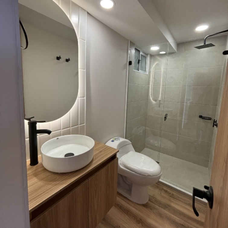 Baño moderno con tocador de madera, lavabo redondo, inodoro y ducha de cristal.