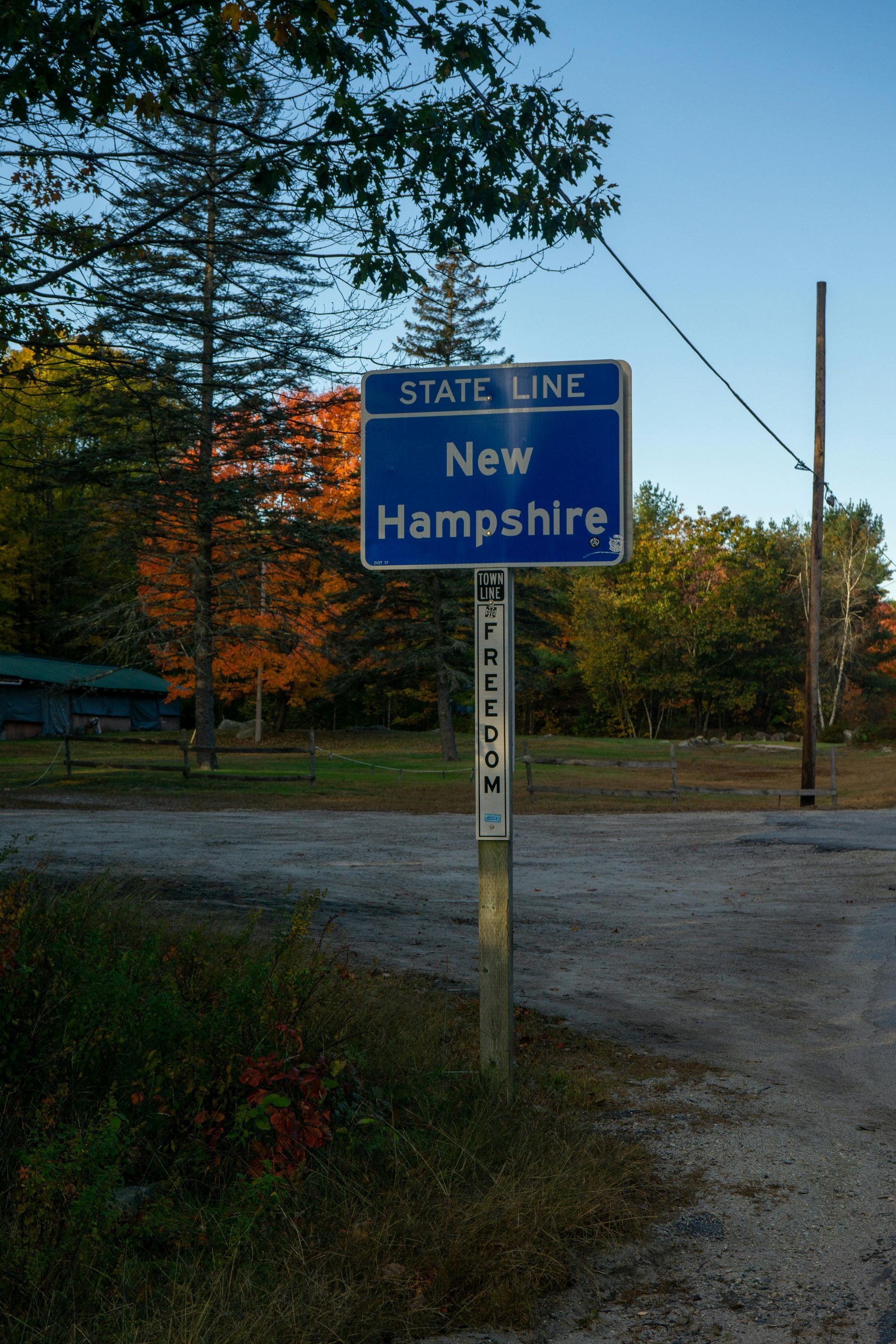 Blue “State Line New Hampshire” sign by a gravel road at dusk, with trees and autumn leaves behind it
