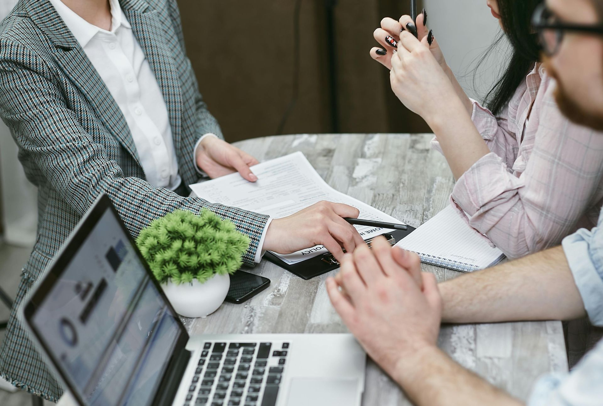 Three people review documents at a table. A laptop and a small plant are present.