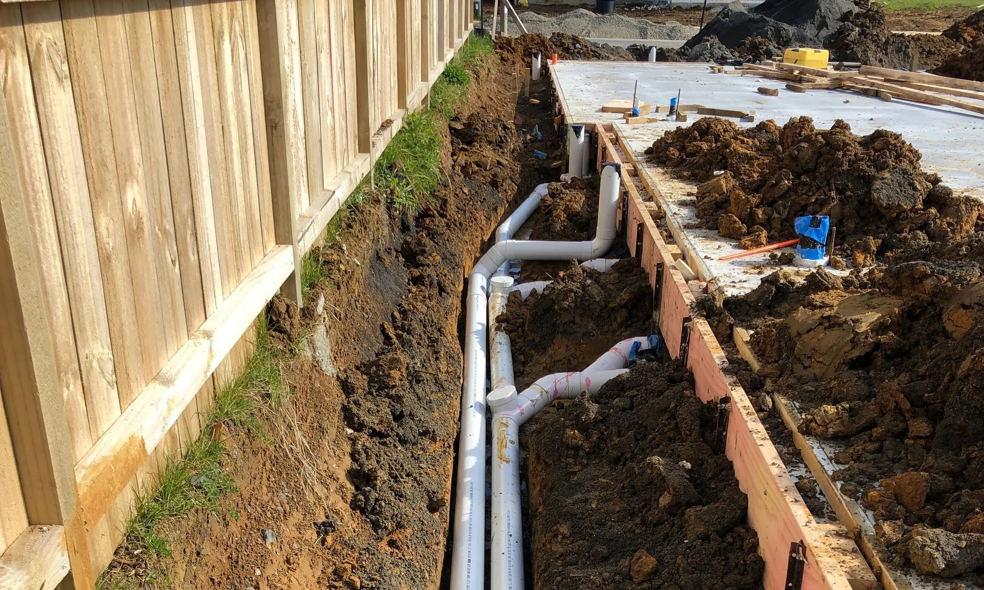 A bunch of pipes are laying in the dirt next to a wooden fence.
