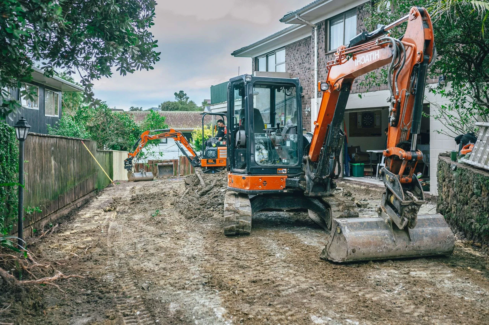 Two excavators are working on a dirt road in front of a house.