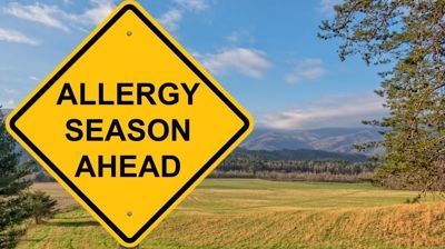 A yellow sign that says `` allergy season ahead '' is in front of a field.