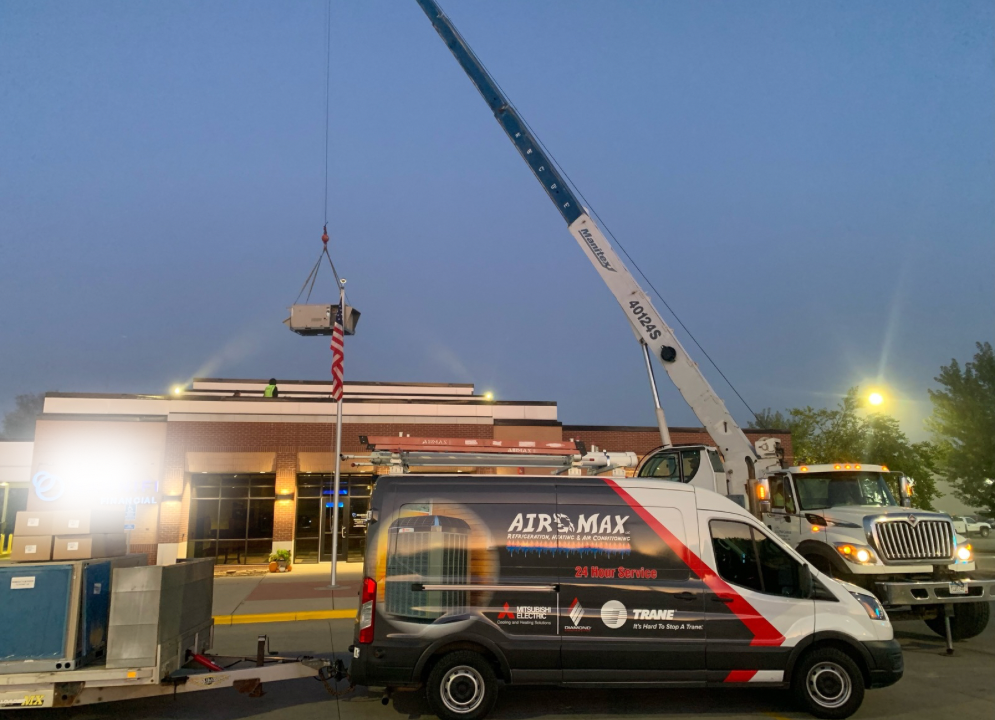 A crane lifts an AC unit onto a building. A service van with 