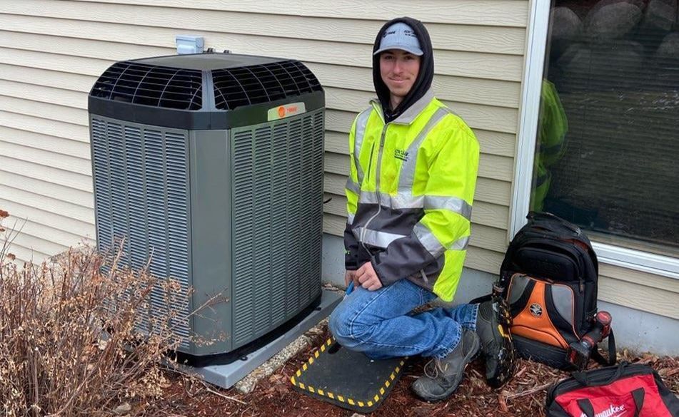 A man is kneeling next to an air conditioner on the side of a house.