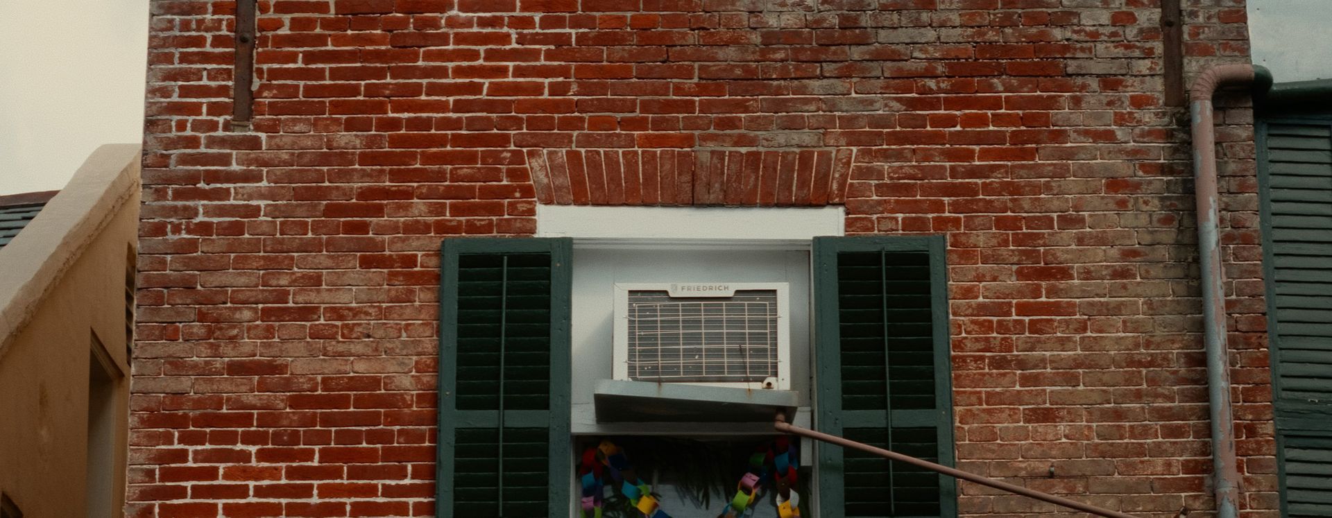 Red brick building with a window featuring an air conditioner and green shutters.