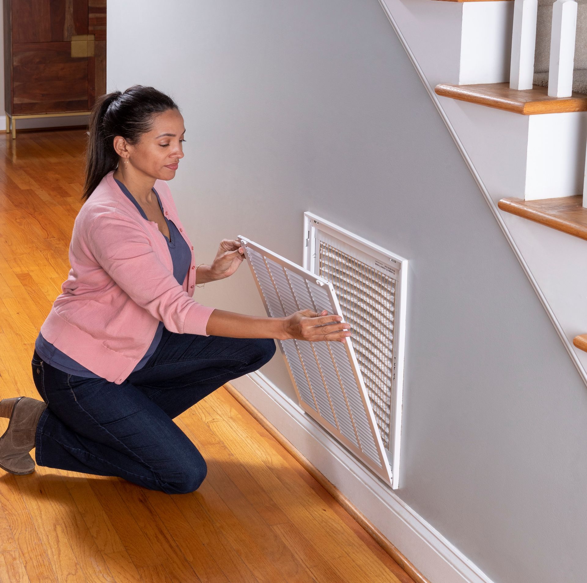 A woman is kneeling down to remove a filter from a wall under a staircase.