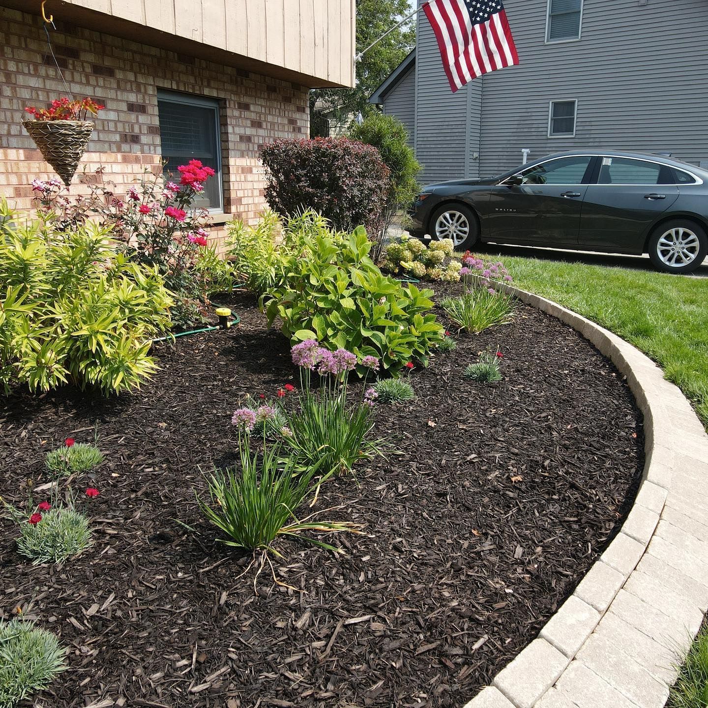 A landscaped garden bed with dark mulch, various plants, and a stone border, next to a brick house and car.