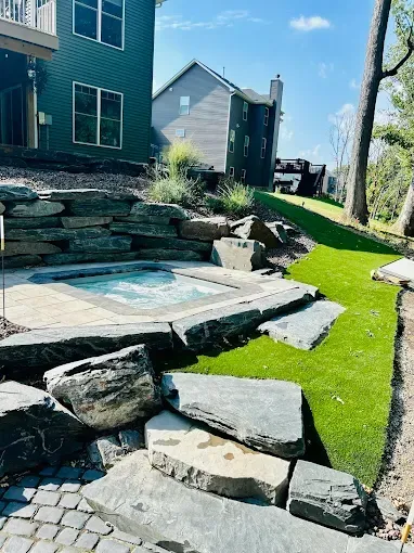 Hot tub surrounded by stone and green grass. Two-story houses in background. Sunny day.
