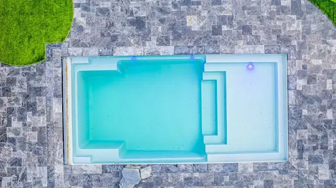 Overhead view of a turquoise rectangular pool with a step entry, surrounded by stone tiles and a patch of green grass.