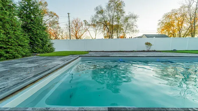 Swimming pool with clear turquoise water, surrounded by a concrete deck and white fence. Green trees and clear sky.