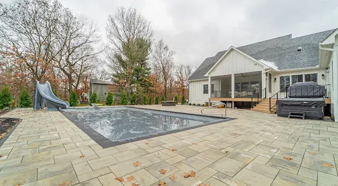 Backyard with pool, slide, patio, and a white house with a covered deck. Overcast day.