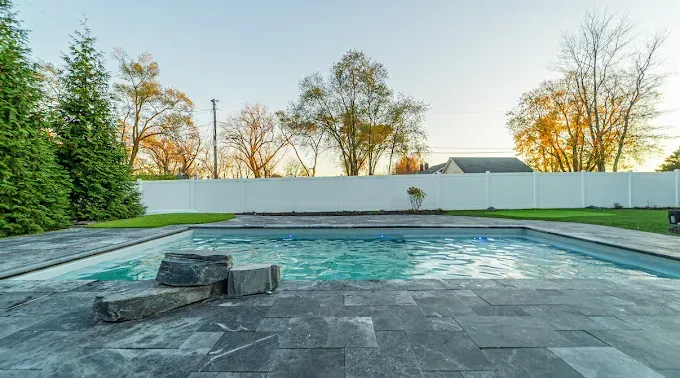 Rectangular pool with stone patio, rocks, and a white fence with trees behind it.