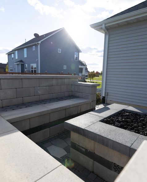Outdoor patio with stone walls, fire pit, bench, and a house in the background on a sunny day.