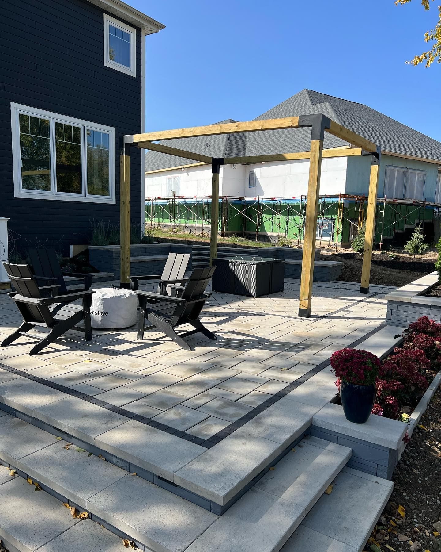 Outdoor patio with gray pavers, wooden pergola, black chairs, and dark blue house.