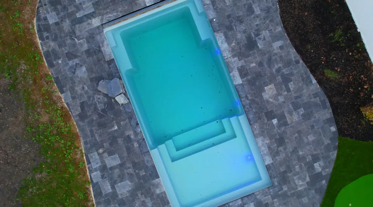 Aerial view of a rectangular pool with blue water, surrounded by gray paving stones, and green grass.