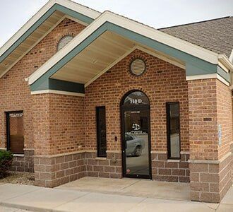 Brick building with a glass door, surrounded by windows. Green trim accents the roof.