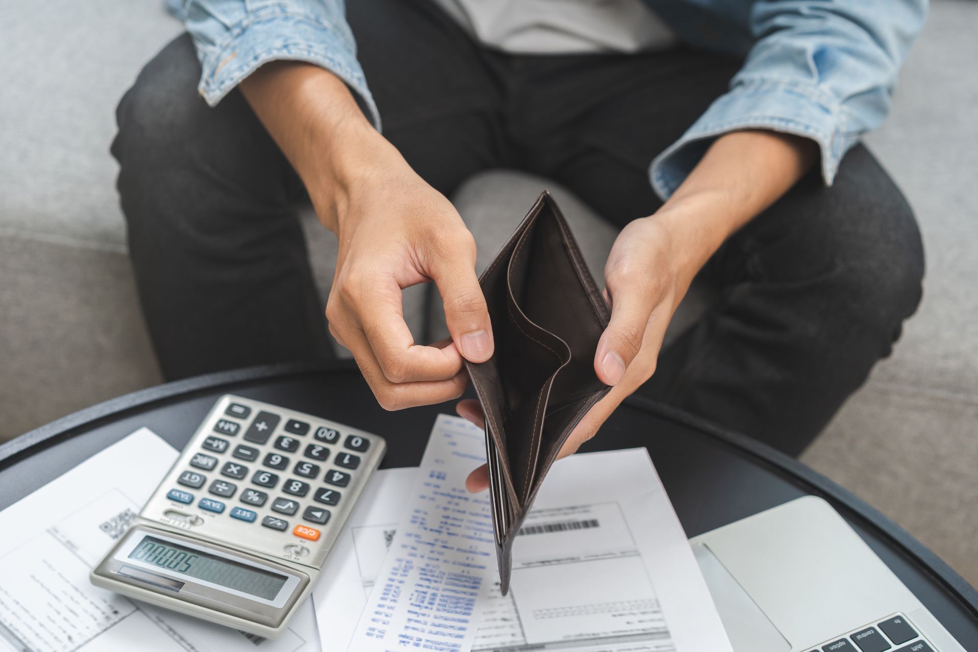 Person holding empty wallet, sitting near calculator, receipts, and laptop; indicating financial difficulties.