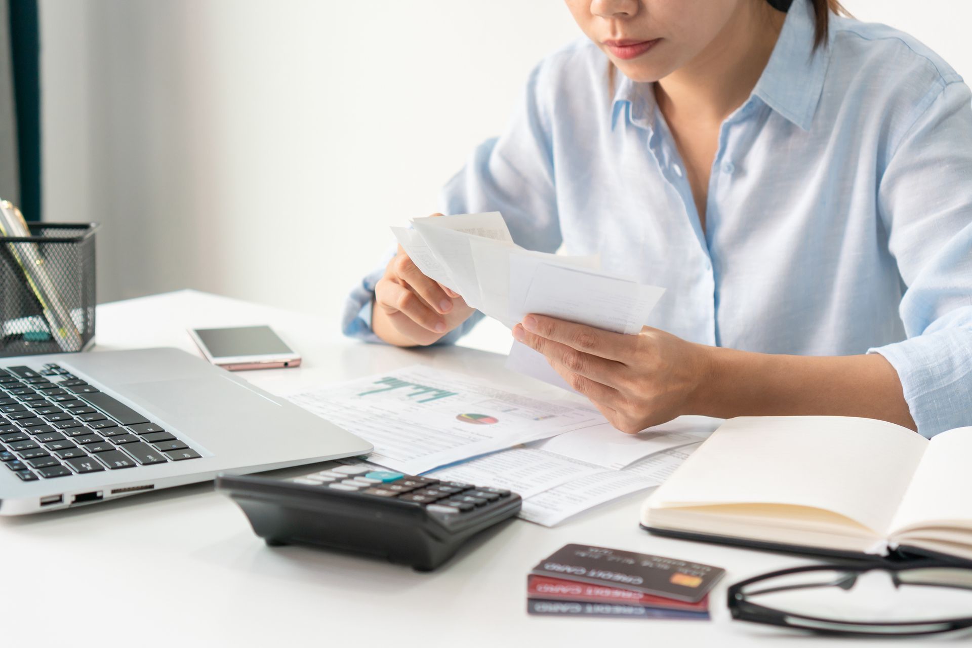 A woman sitting at her desk is calculating monthly expenses at home with a laptop, notebook, and cal