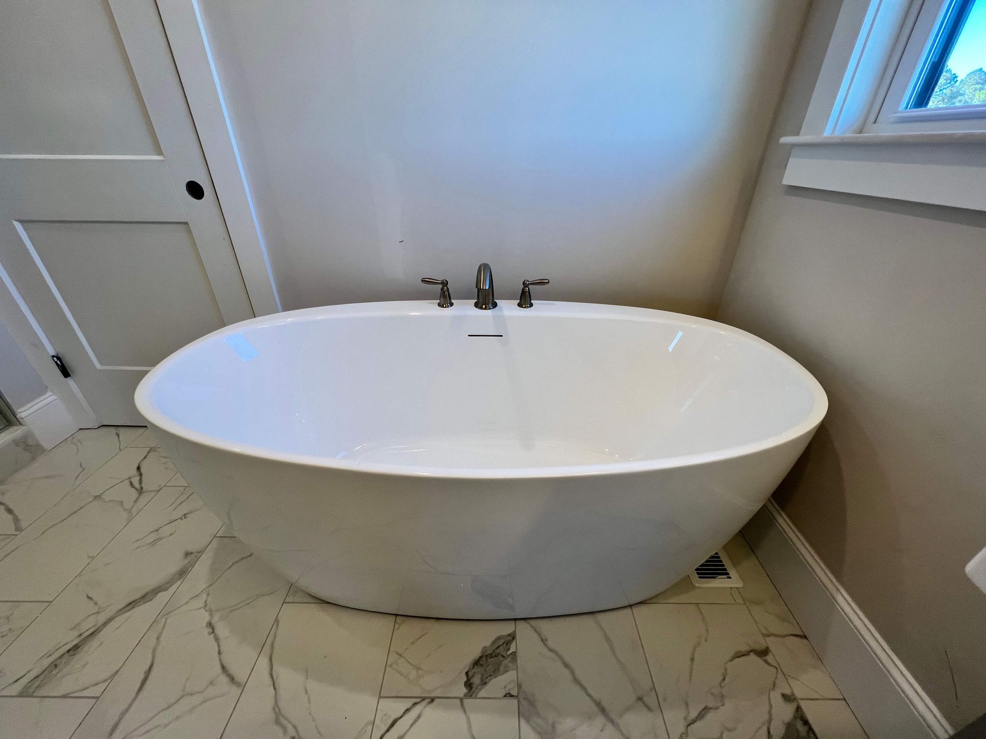 White oval freestanding bathtub in a bathroom with marble tile floor and window.