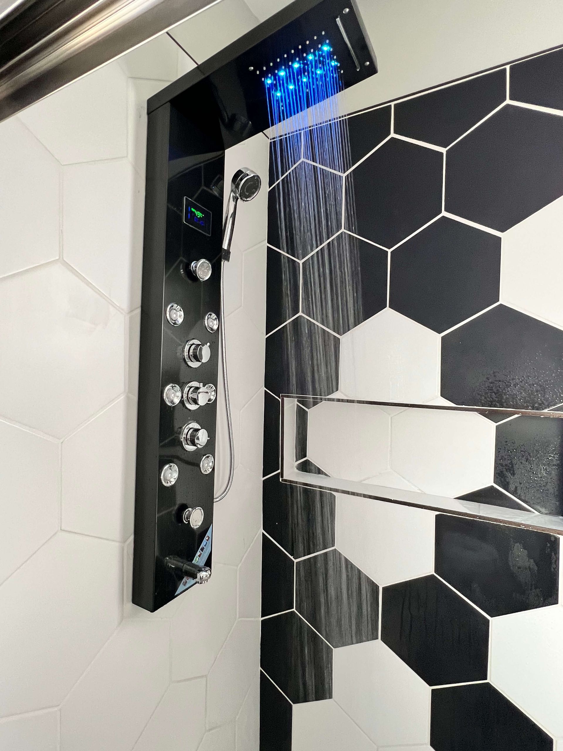 Black shower panel with blue overhead lights, next to hexagonal black and white tiled wall.