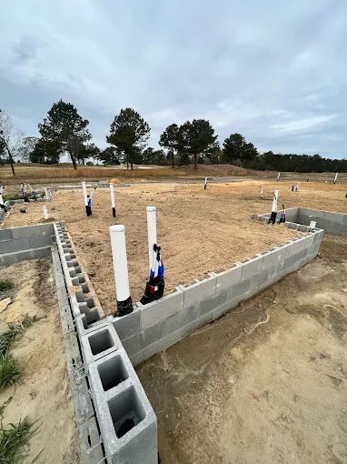 Foundation of a building under construction. Concrete block walls, white pipes, and a dirt base. Cloudy sky in the background.