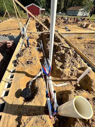Construction site with PVC pipes and blue/red water lines in a trench next to cinder blocks and wooden framing.