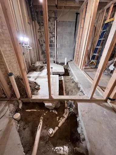 Bathroom renovation in progress, framing and plumbing visible. Exposed pipes and concrete floor.