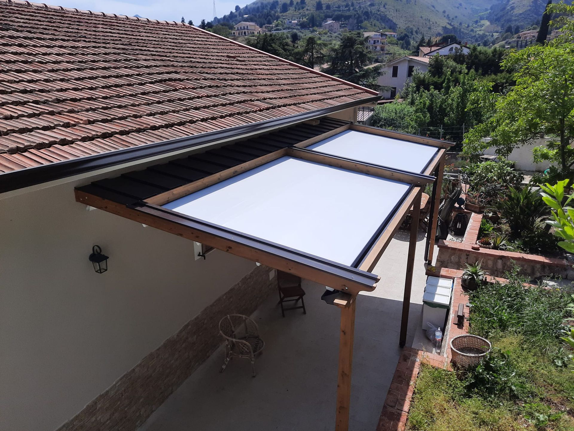 Patio con tenda da sole bianca retrattile sotto un tetto di tegole, pali di legno e vista sugli alberi.