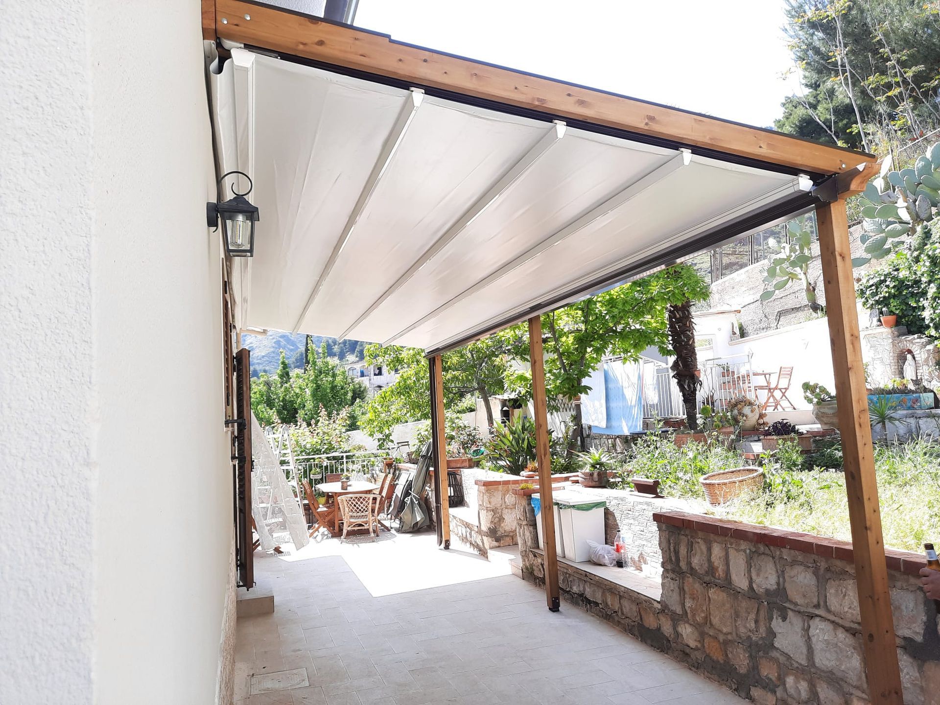 Pergola in legno con tettoia bianca retrattile sopra il patio, adiacente a un edificio.